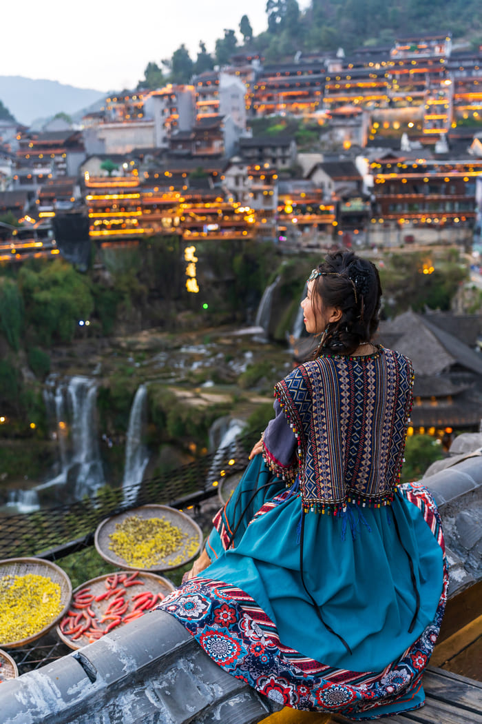 Viajera con vestimenta tradicional observando un antiguo pueblo histórico iluminado junto a una cascada en China